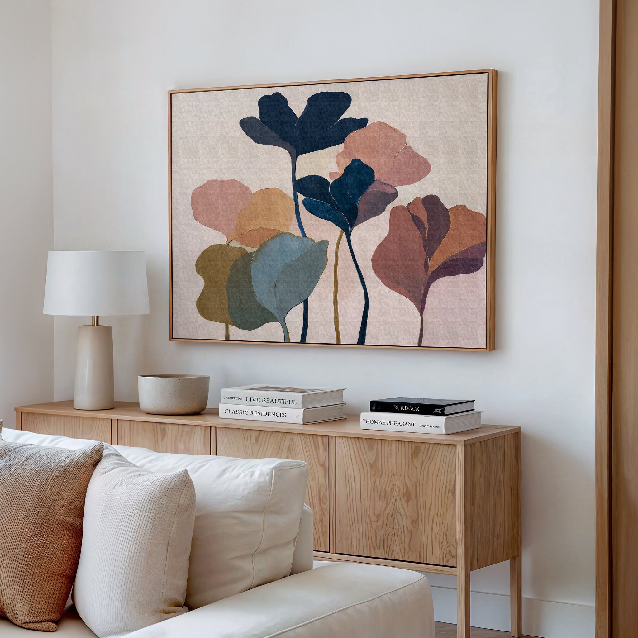 A modern living room features a wooden sideboard, beige and white cushions, a table lamp, and stacked books. Above the sideboard hangs the Autumn Garden Canvas Art - Horizontal, adding vibrant autumn flair as striking modern wall art.