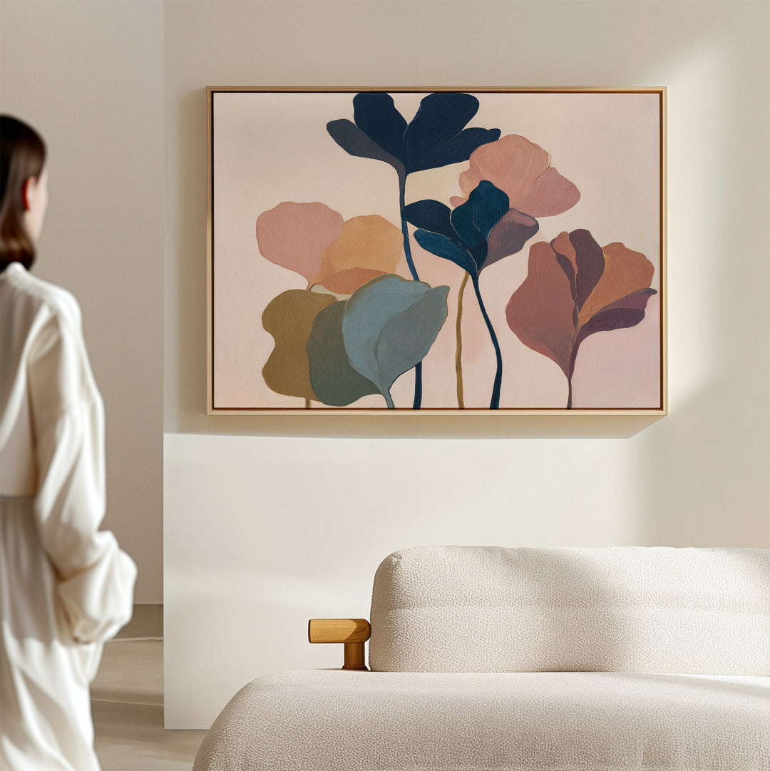 A woman in a white outfit stands in a bright, minimal room, admiring the "Autumn Garden Canvas Art - Horizontal," featuring colorful, overlapping leaf shapes displayed above a beige textured sofa.