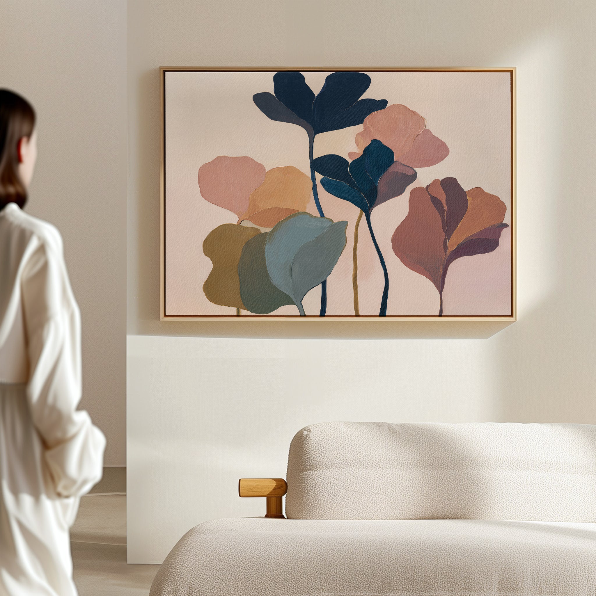 A woman in a white outfit stands in a bright, minimal room, admiring the "Autumn Garden Canvas Art - Horizontal," featuring colorful, overlapping leaf shapes displayed above a beige textured sofa.