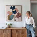 A woman in a white blouse and jeans leans against a wooden cabinet, gazing at the Autumn Garden Canvas Art - Square—a modern botanical piece that brings vibrant color to the white wall above, with books and a small potted plant completing the decor.