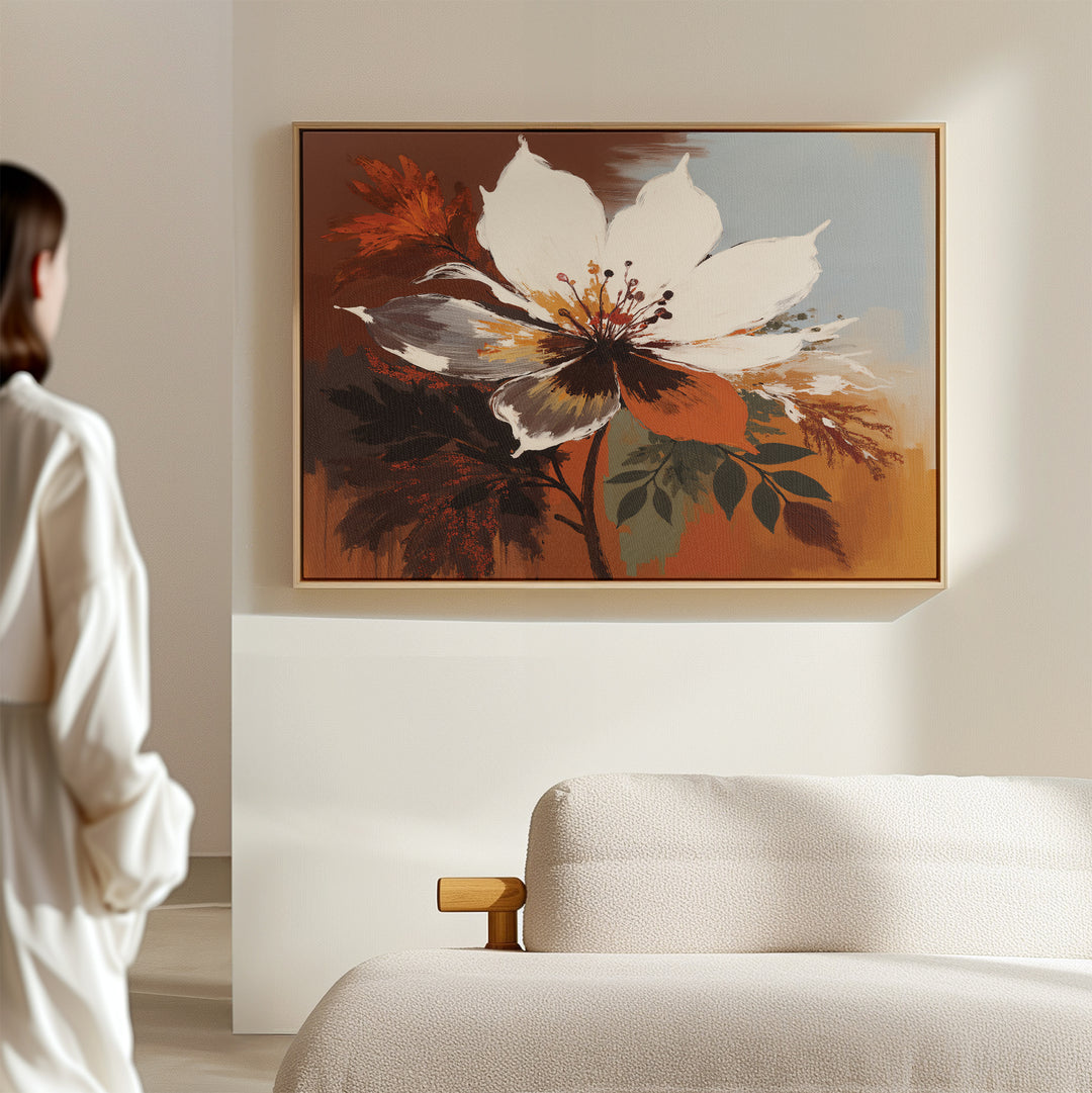 A person in a white outfit admires the Autumn Radiance Canvas Art - Horizontal, a bold floral print with warm brown, orange, and cream tones, displayed above a textured beige sofa in a modern minimal living room.