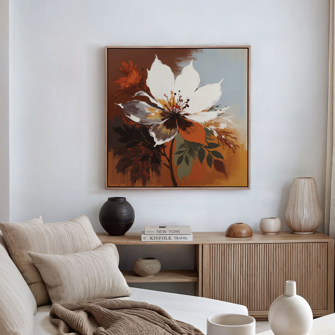 A cozy living room with a beige sofa, neutral-toned cushions, and a wooden sideboard topped with vases and books. Above it hangs the Autumn Radiance Canvas Art - Square, featuring bold white, orange, and brown florals.