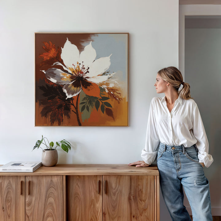 A woman in a white blouse and jeans stands by a wooden cabinet, admiring the Autumn Radiance Canvas Art - Square on the wall. A potted plant and a book rest on the cabinet, highlighting the room’s minimalist, modern style.