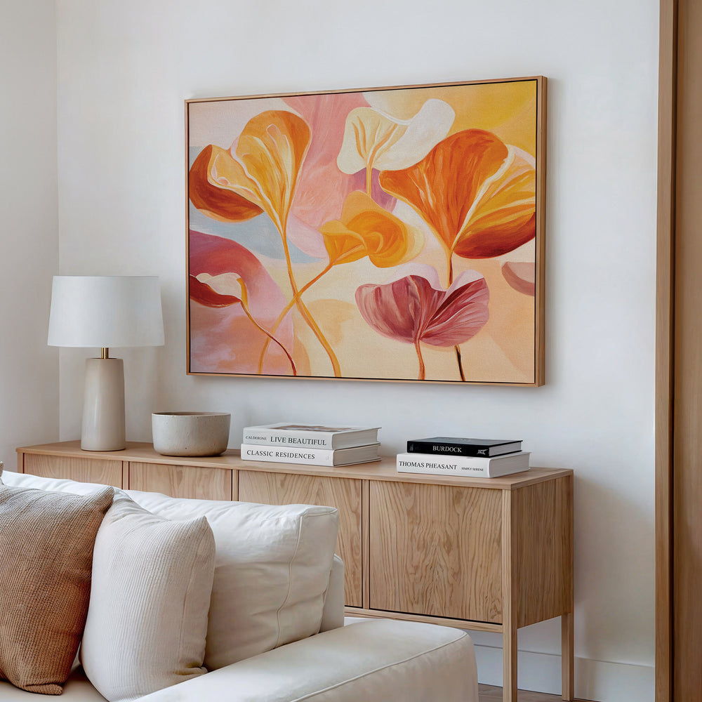 A cozy living space features a wooden sideboard with stacked books, a white lamp, and bowl. Above hangs the Autumn Reverie Canvas Art - Horizontal in warm tones, while a cushioned sofa sits in the foreground.