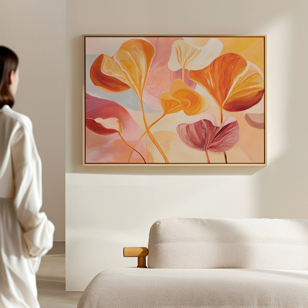 A person in a white shirt admires the Autumn Reverie Canvas Art - Horizontal, featuring orange, yellow, and pink leaves above a beige sofa in a softly lit minimalist room.