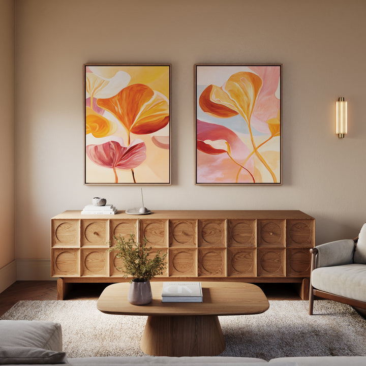 A cozy living room features a wooden sideboard, coffee table, sofa, and "Autumn Reverie - Set of 2" botanical wall art. A vase with flowers sits on the table as a wall sconce casts warm light, completing the floral decor.