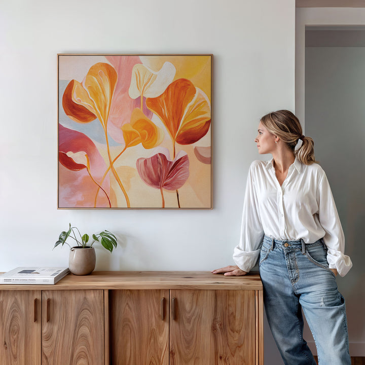 A woman in a white blouse and jeans leans against a wooden cabinet, admiring the Autumn Reverie Canvas Art - Square, a colorful abstract floral piece above her. A small plant and books on the cabinet complete the cozy fall floral decor.