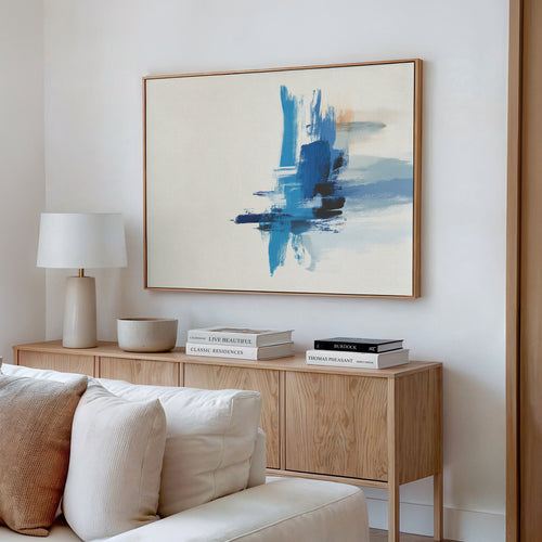 A modern living room features a light wooden sideboard, a white table lamp, stacked books, and the Azure Convergence Canvas Art - Horizontal in blue tones displayed above a white sofa with cream cushions.