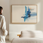 A person admires the Azure Convergence Canvas Art - Horizontal, featuring blue and orange brushstrokes, displayed on a light wall above a textured beige sofa in a modern, minimalist room.