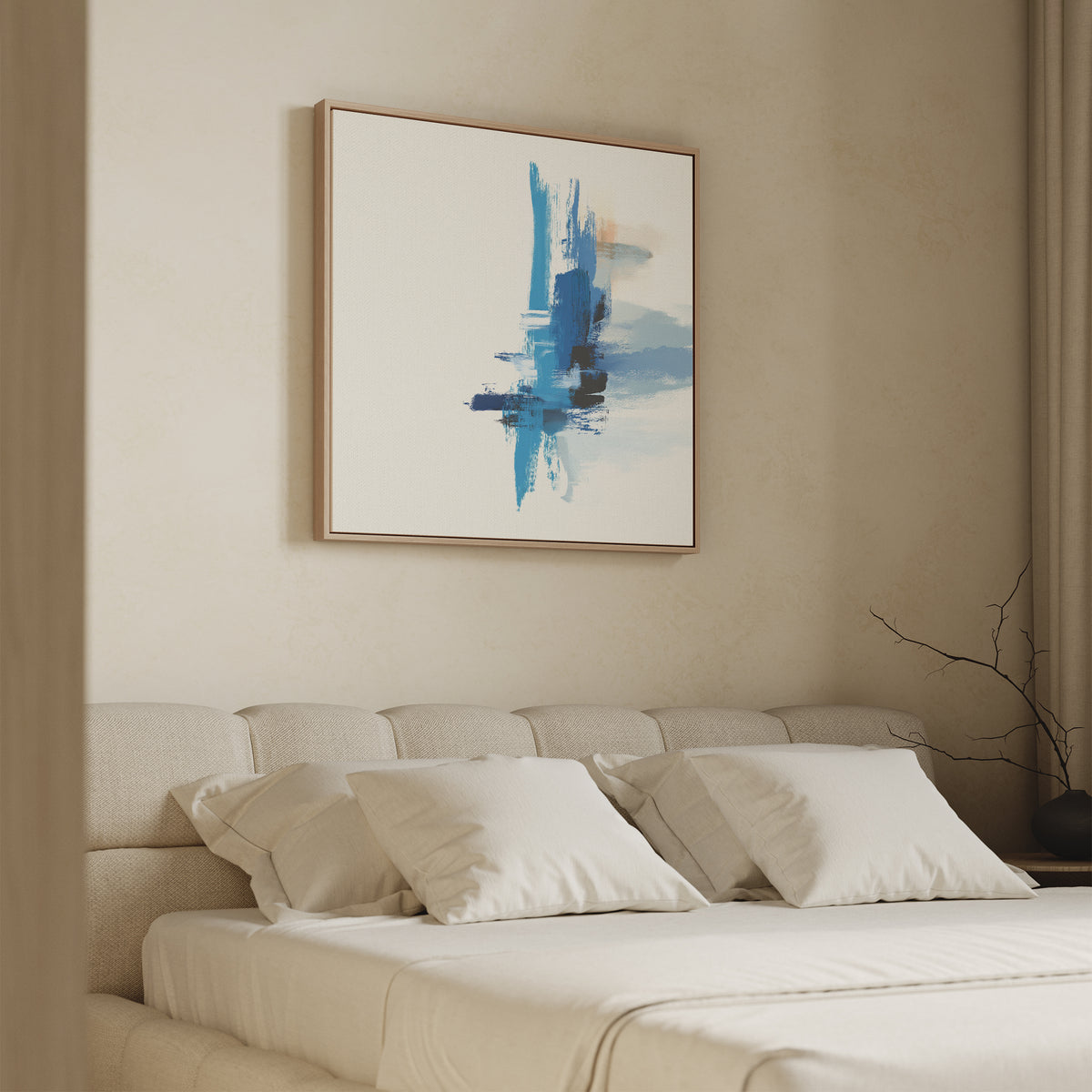 A beige bedroom features a cushioned headboard, white bedding and pillows. The Azure Convergence Canvas Art - Square in blue and white hangs above the bed. A bare branch decorates a nightstand near drawn beige curtains.