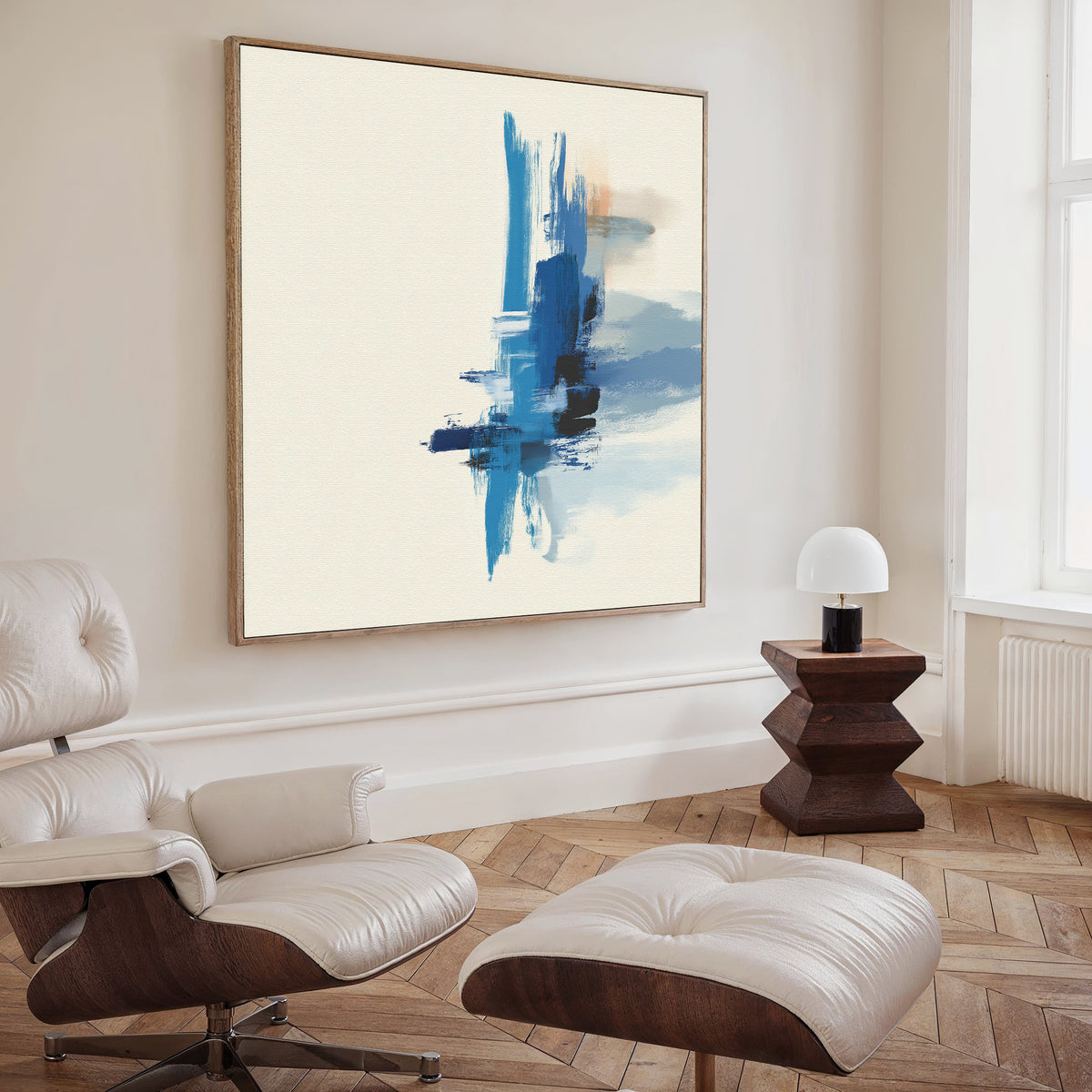 A modern living room with a white lounge chair and ottoman, a wooden side table with a small white lamp, and the Azure Convergence Canvas Art - Square—a bold abstract painting featuring blue brushstrokes on a pale background.