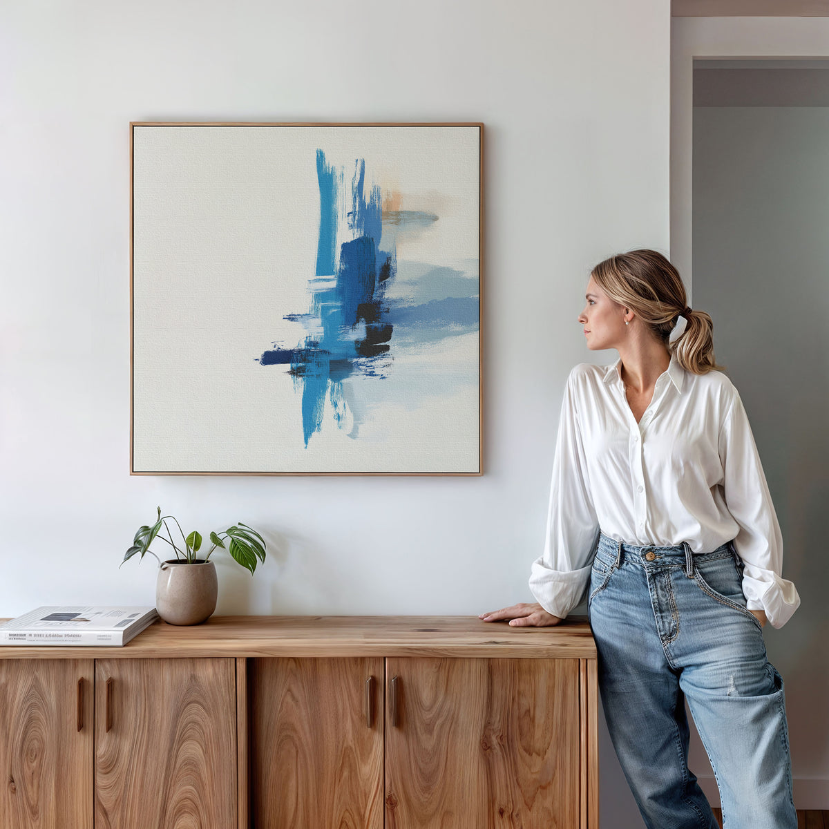 A woman in a white blouse and jeans leans against a wooden cabinet topped with books and a potted plant, gazing at the Azure Convergence Canvas Art - Square, a modern blue and white abstract print hung above her.