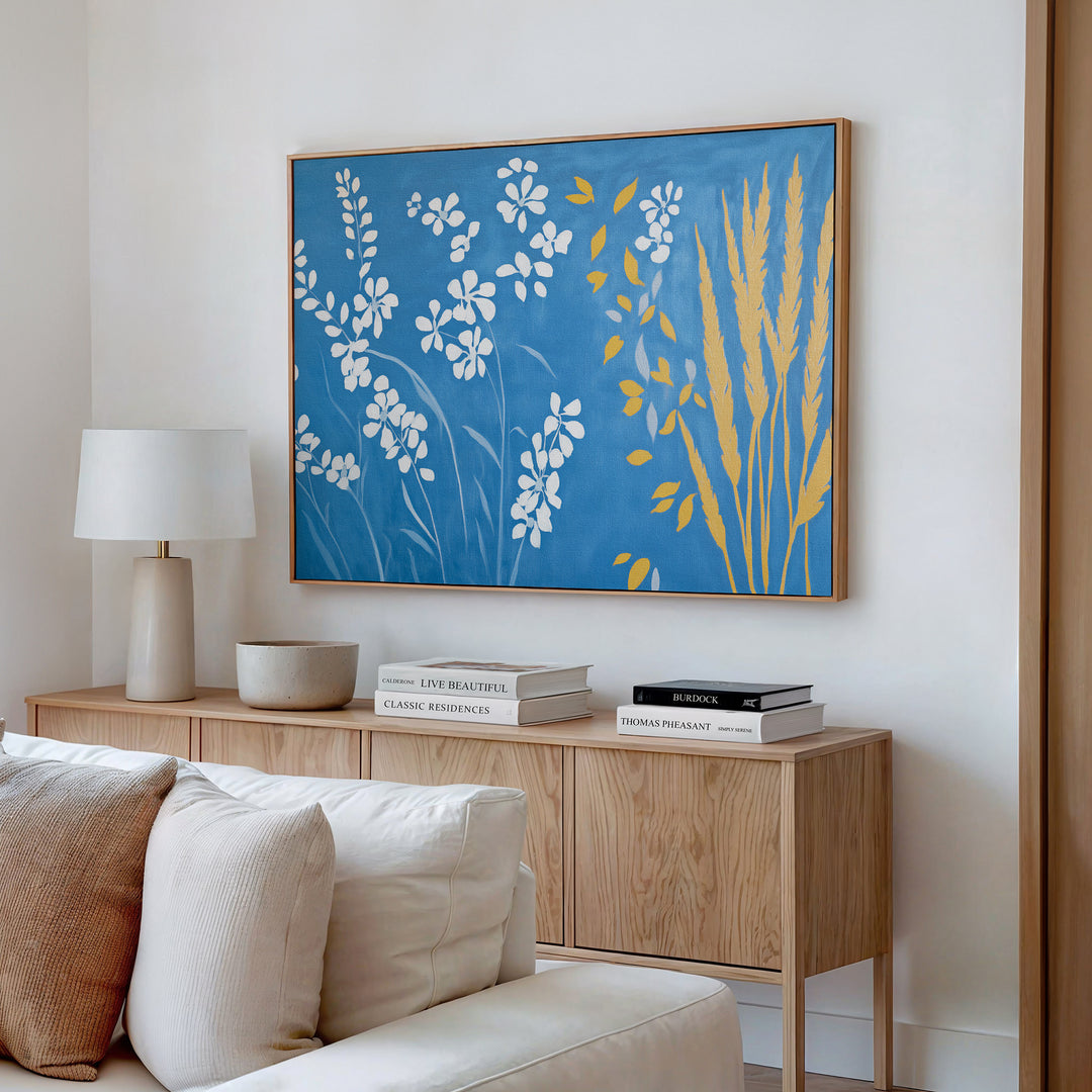 A modern living room showcases a light wood console table with stacked books, a lamp, and the Azure Meadow Canvas Art - Horizontal—a large botanical piece in blue, white, and yellow—hung above it. A beige sofa with plush cushions sits in front.