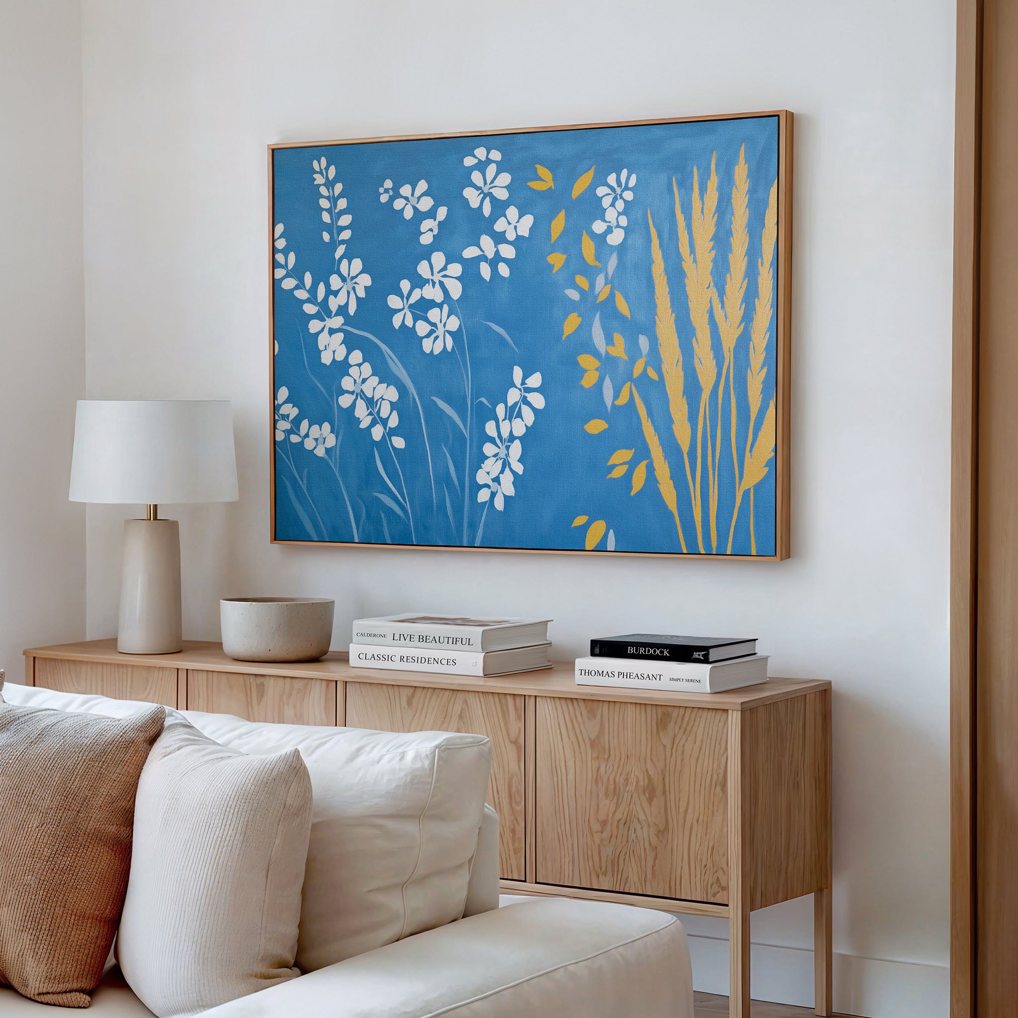 A modern living room showcases a light wood console table with stacked books, a lamp, and the Azure Meadow Canvas Art - Horizontal—a large botanical piece in blue, white, and yellow—hung above it. A beige sofa with plush cushions sits in front.