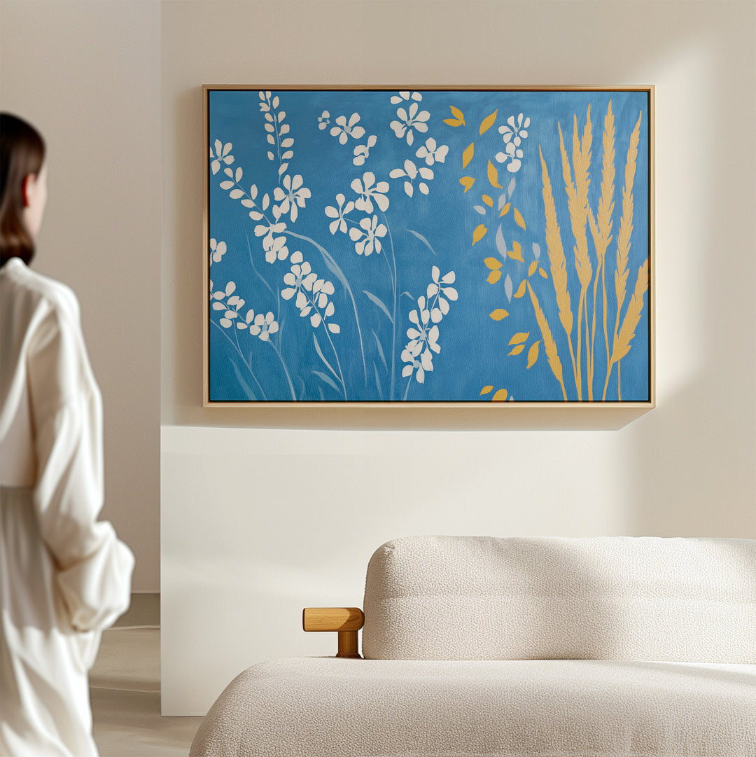 A person stands in a minimal living room, admiring the Azure Meadow Canvas Art - Horizontal, which features white and yellow botanical shapes on a blue background above a light beige sofa.