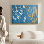 A person stands in a minimal living room, admiring the Azure Meadow Canvas Art - Horizontal, which features white and yellow botanical shapes on a blue background above a light beige sofa.
