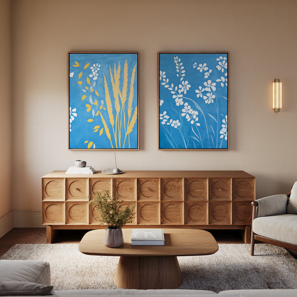 A modern living room features a wooden sideboard, light wood coffee table, and beige sofa. Above the sideboard, the Azure Meadow - Set of 2 prints add color, while white wildflowers in a vase decorate the table.