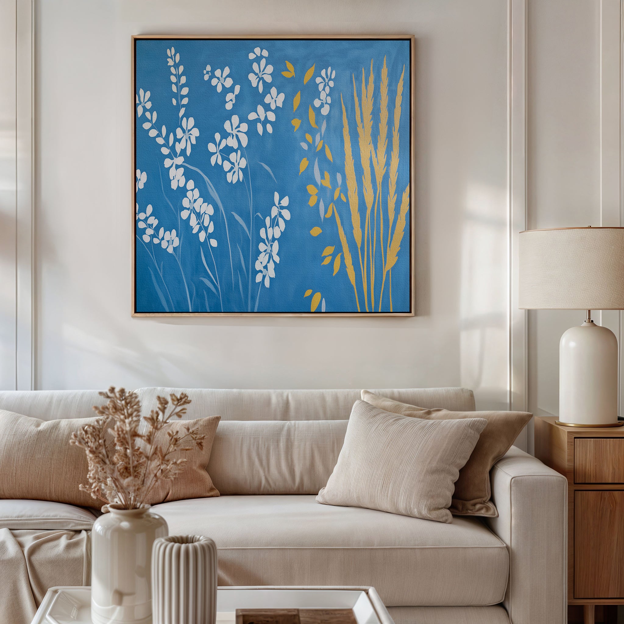 A modern living room featuring a white sofa, beige pillows, a side table with a lamp, and a vase of dried flowers. Above the sofa hangs Azure Meadow Canvas Art - Square, showcasing striking blue botanical designs with white and yellow flowers.