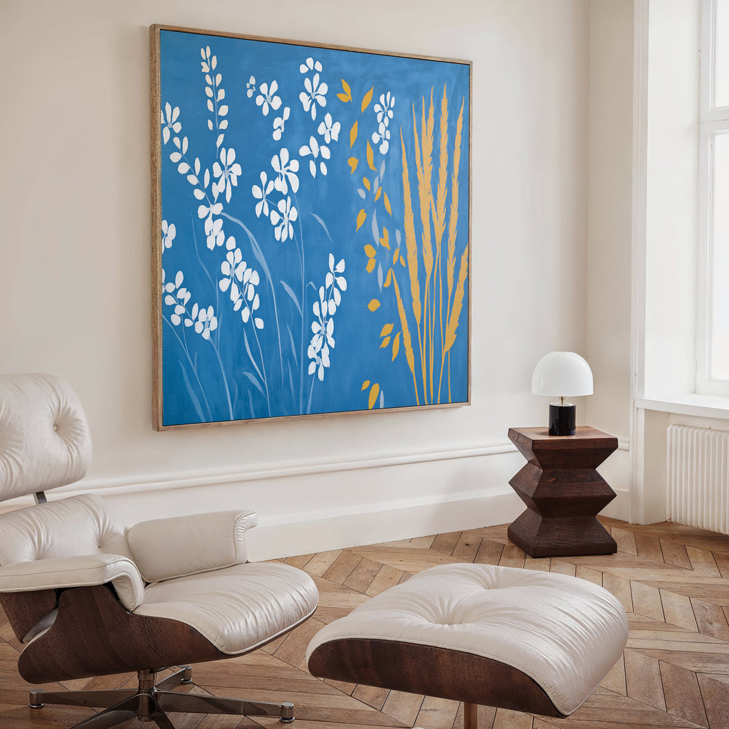 A modern living room featuring a white lounge chair and ottoman, wooden side table with a white lamp, and Azure Meadow Canvas Art - Square, showcasing blue, white, and yellow floral shapes. Light herringbone floor and bright window.