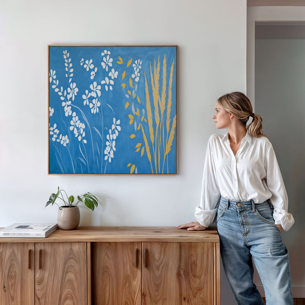 A woman in a white blouse and jeans leans on a wooden cabinet, admiring the Azure Meadow Canvas Art - Square. A small plant and books are arranged on the cabinet, complementing the botanical wall art.