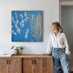 A woman in a white blouse and jeans leans on a wooden cabinet, admiring the Azure Meadow Canvas Art - Square. A small plant and books are arranged on the cabinet, complementing the botanical wall art.