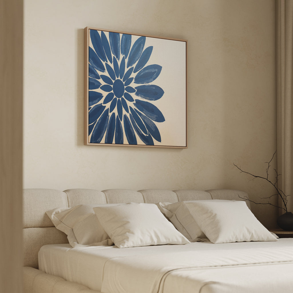 A minimalist bedroom features a beige upholstered bed with white bedding and pillows. Above, the Azure Radiance Canvas Art - Square showcases a bold blue flower. Soft natural light and neutral walls complete the calm, modern decor.