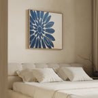 A minimalist bedroom features a beige upholstered bed with white bedding and pillows. Above, the Azure Radiance Canvas Art - Square showcases a bold blue flower. Soft natural light and neutral walls complete the calm, modern decor.
