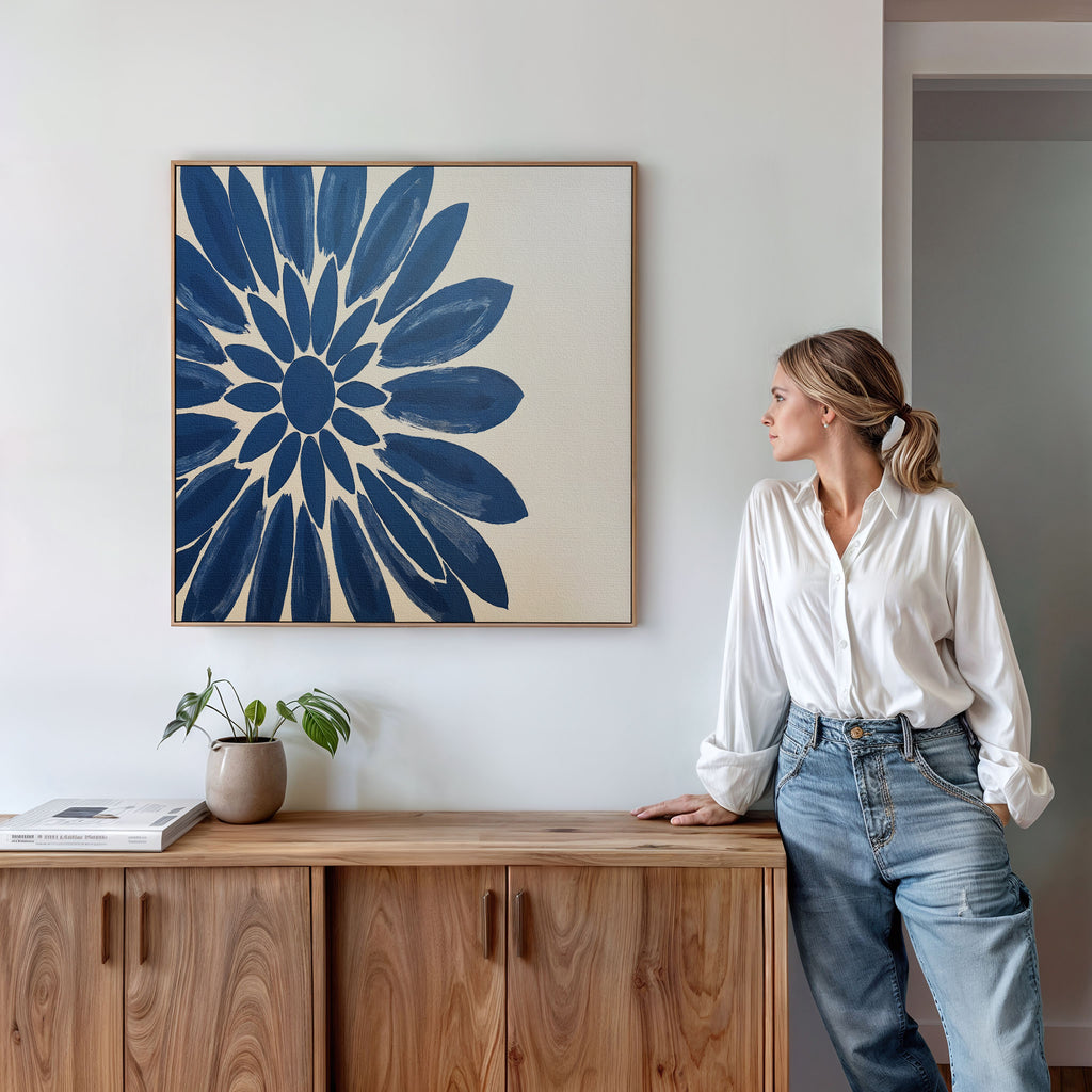 A woman in a white blouse and jeans leans on a wooden cabinet, gazing at the "Azure Radiance Canvas Art - Square" above. A small plant and book rest on the cabinet in this modern, minimalist room—ideal for home decor inspiration.