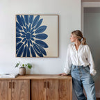 A woman in a white blouse and jeans leans on a wooden cabinet, gazing at the "Azure Radiance Canvas Art - Square" above. A small plant and book rest on the cabinet in this modern, minimalist room—ideal for home decor inspiration.