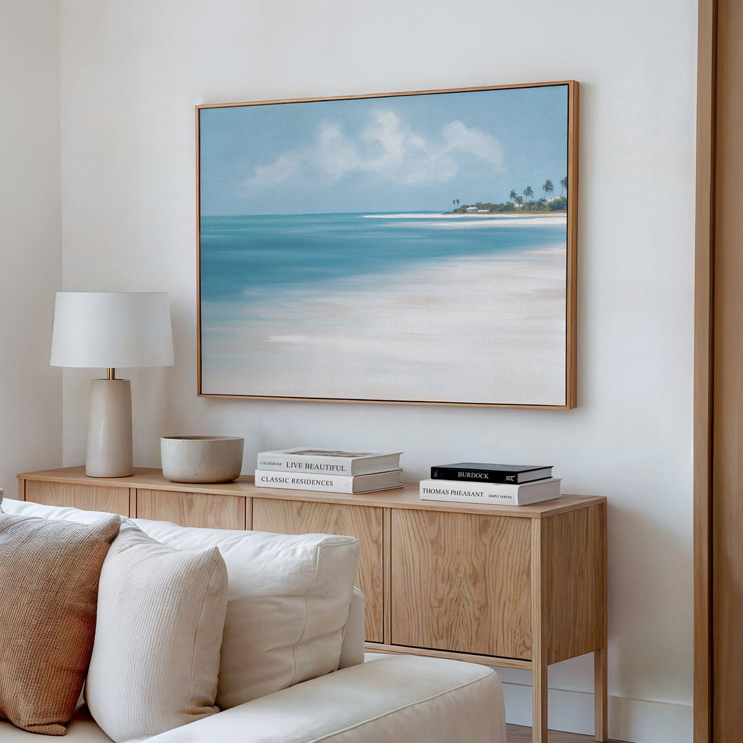 A bright, modern living room showcases a wooden sideboard with books, a white lamp, and a bowl. Above it hangs the Azure Tranquility Canvas Art - Horizontal, featuring turquoise waters. A light-colored sofa is partly visible in front.