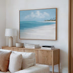A bright, modern living room showcases a wooden sideboard with books, a white lamp, and a bowl. Above it hangs the Azure Tranquility Canvas Art - Horizontal, featuring turquoise waters. A light-colored sofa is partly visible in front.
