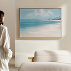 A woman stands in a softly lit room, gazing at the Azure Tranquility Canvas Art - Horizontal, featuring serene turquoise waters and distant palm trees, displayed on a light-colored wall above a beige sofa.
