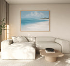 A modern living room features a large cream sectional, a light wood coffee table, and the Azure Tranquility Canvas Art - Horizontal depicting turquoise waters. Neutral tones and natural light create a calm, minimalist vibe.