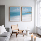 Minimalist living room with a light gray sofa, woven chair, and small round table. The "Azure Tranquility - Set of 2" wall art featuring turquoise waters hangs above the chair, while white candles and large windows create a calm, naturally lit space.