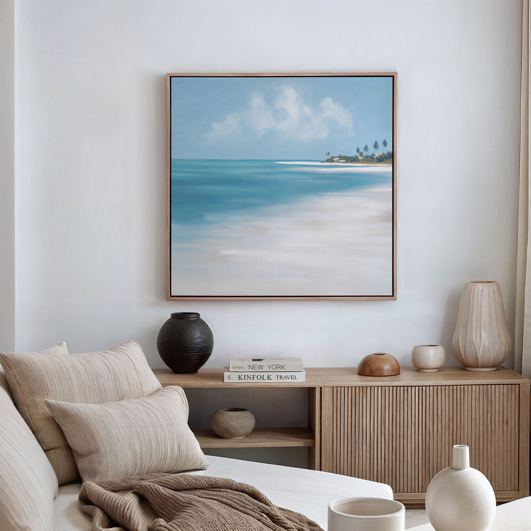 A modern, minimalist living room with a beige sofa, neutral cushions, wooden sideboard, vases, and serene Azure Tranquility Canvas Art - Square featuring a beach landscape hanging on a white wall.