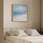 A minimalist bedroom features a beige upholstered bed with cream bedding. Above, the Azure Tranquility Canvas Art - Square depicts blue water, white sand, and palm trees, bringing a calming coastal vibe to the space.