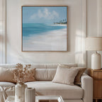 A cozy living room with a beige sofa, neutral pillows, and a side table with a lamp is accented by vases of dried flowers. The Azure Tranquility Canvas Art - Square hangs above the sofa, adding relaxing coastal vibes to the space.