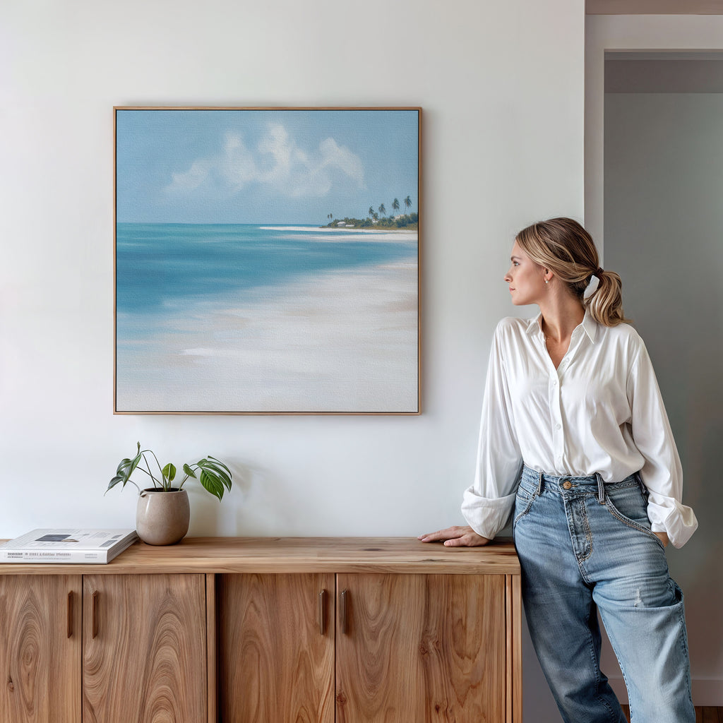 A woman in a white blouse and jeans leans against a wooden sideboard, admiring the Azure Tranquility Canvas Art - Square. The serene turquoise waters and beach scene, paired with a small plant and book, create a peaceful setting.