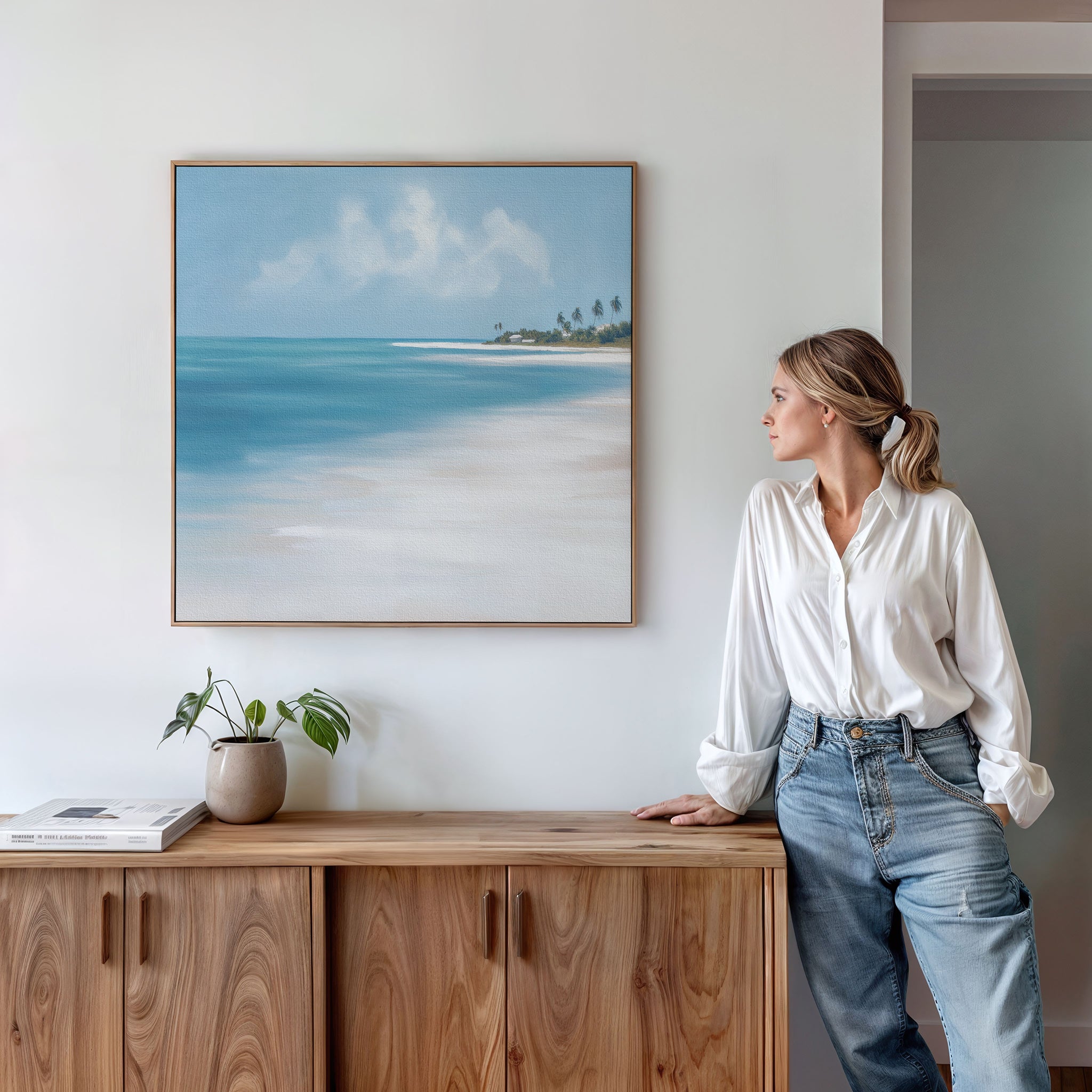 A woman in a white blouse and jeans leans against a wooden sideboard, admiring the Azure Tranquility Canvas Art - Square. The serene turquoise waters and beach scene, paired with a small plant and book, create a peaceful setting.