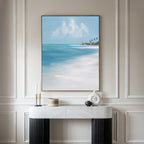 The Azure Tranquility Canvas Art - Vertical hangs on a cream wall above a marble console, its beach scene with turquoise waters complemented by minimalist decor—candles, a wooden box, and a white abstract sculpture—for a serene display.