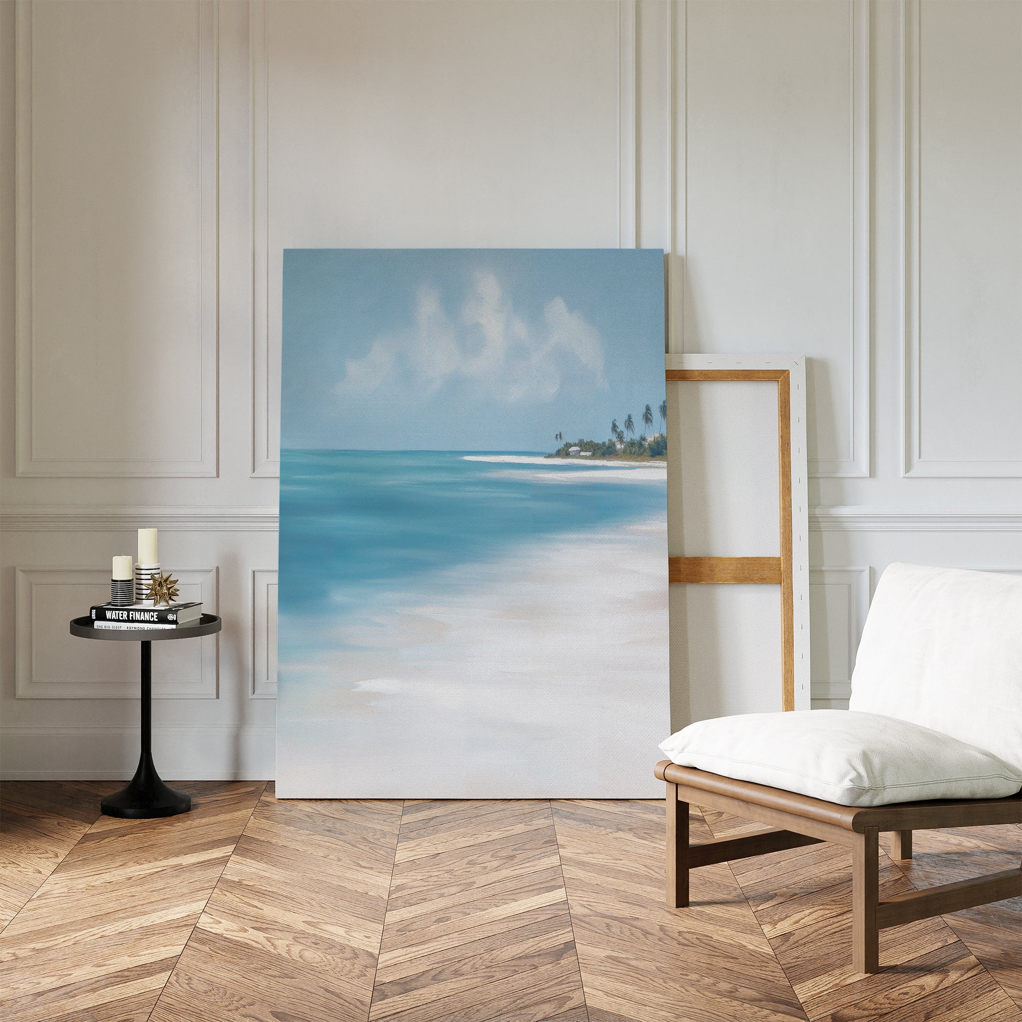 The Azure Tranquility Canvas Art - Vertical, featuring serene beach scenery and turquoise waters, rests against a white paneled wall on herringbone wood flooring beside a cushioned chair, empty wooden frame, and small round table with candles and books.