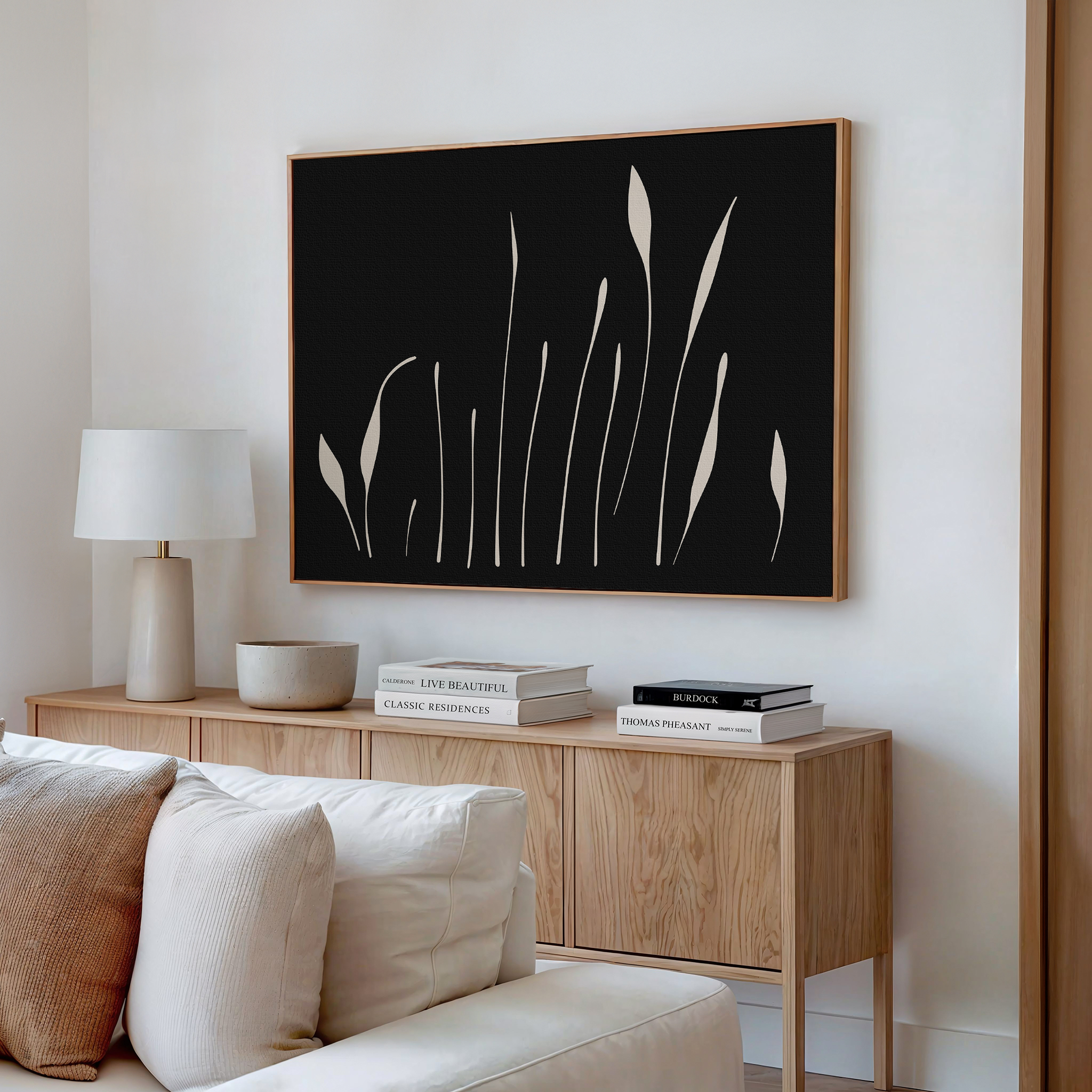A modern living room features a light wood sideboard, white table lamp, ceramic bowl, stacked books, and Botanical Silhouettes Abstract Canvas Art with plant-like shapes above. A cream-colored sofa is in the foreground.
