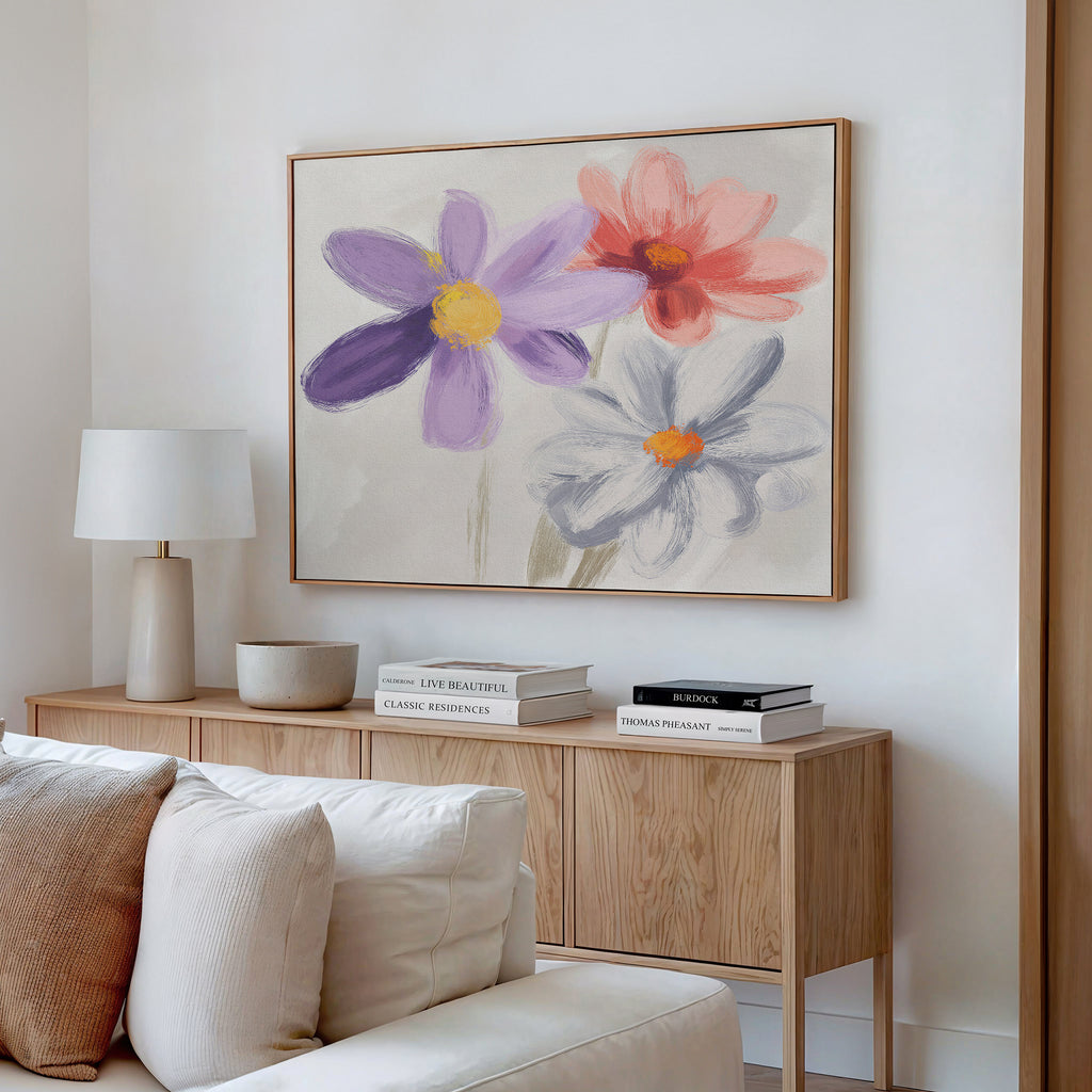 A cozy living room features a light wood cabinet, books, a modern lamp, and the Blooming Harmony Canvas Art - Horizontal with vibrant purple, red, and white flowers above. Soft neutrals evoke a calm Nordic Park-inspired atmosphere.