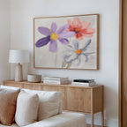 A cozy living room features a light wood cabinet, books, a modern lamp, and the Blooming Harmony Canvas Art - Horizontal with vibrant purple, red, and white flowers above. Soft neutrals evoke a calm Nordic Park-inspired atmosphere.