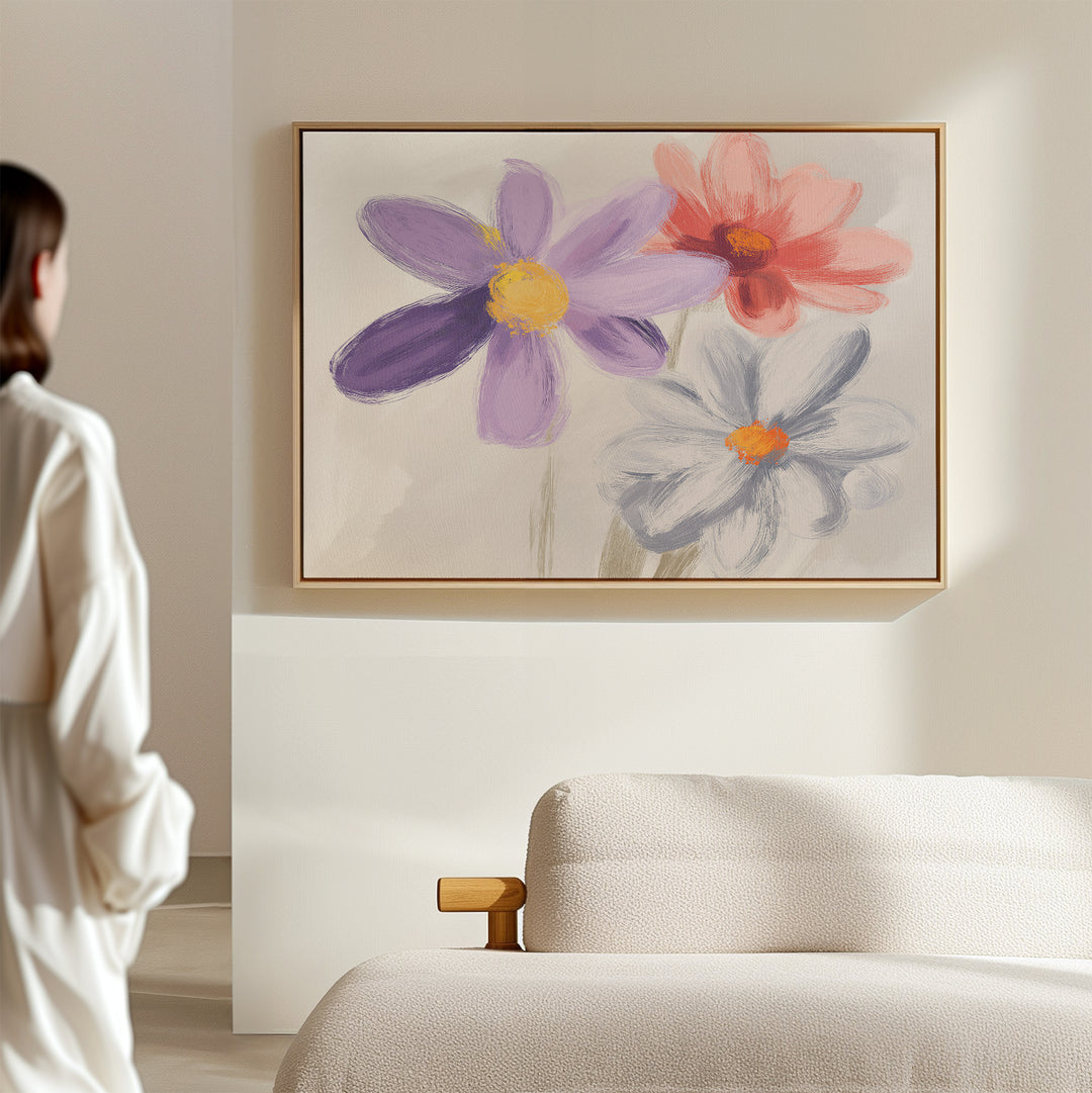 In a softly lit room, a person admires the Blooming Harmony Canvas Art - Horizontal—a vibrant print of purple, pink, and white flowers—displayed above a modern cream-colored sofa.