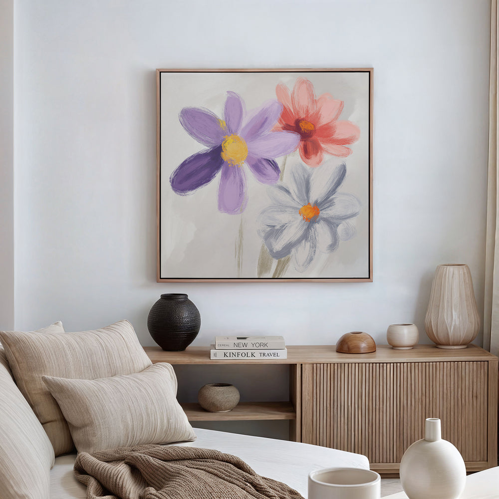 A cozy living room with a beige sofa, neutral cushions, a light wood sideboard, decorative vases, and the Blooming Harmony Canvas Art - Square featuring three colorful flowers. Neatly stacked books on the sideboard complete the botanical decor.