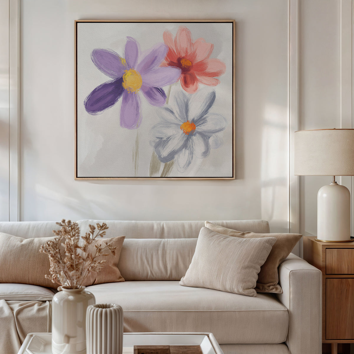A cozy living room with a beige sofa, cushions, a light wood side table with lamp, dried flowers in a vase, and the Blooming Harmony Canvas Art - Square featuring three colorful flowers displayed above the sofa.