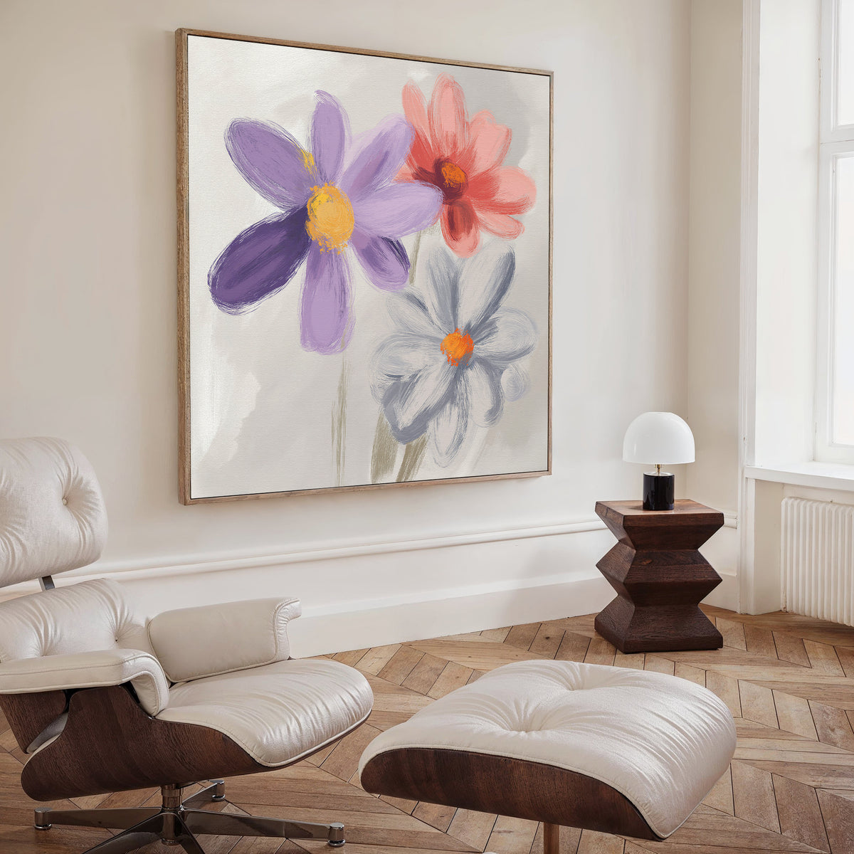 A modern living room showcases the Blooming Harmony Canvas Art - Square, featuring three vibrant flowers, alongside an off-white tufted lounge chair with ottoman, a geometric wooden side table, a lamp, and herringbone wood floors.