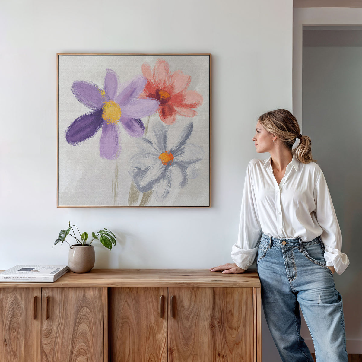 A woman in a white blouse and jeans leans against a wooden cabinet, gazing at the Blooming Harmony Canvas Art - Square hanging above her. A potted plant and books on the cabinet complete the room’s botanical decor.