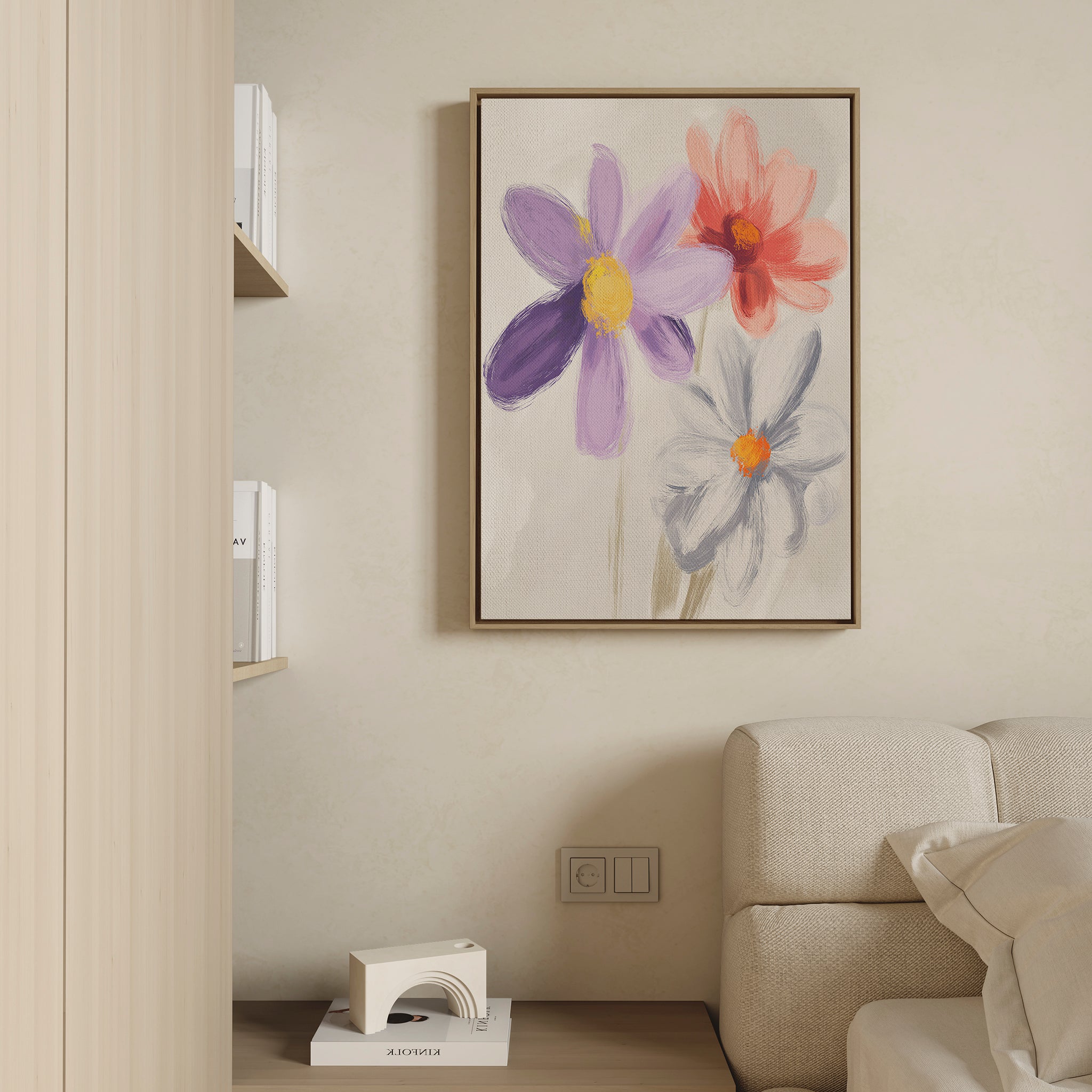 The Blooming Harmony Canvas Art - Vertical, featuring three vibrant flowers, hangs above a cream sofa on a light beige wall in this modern minimalist living room. A small bookshelf sits to the left, enhancing the botanical charm.